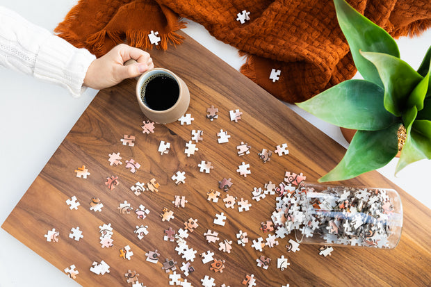 A women holding a coffee mug on a wooden table with the Boobs Puzzle glass jar spilling pieces across.